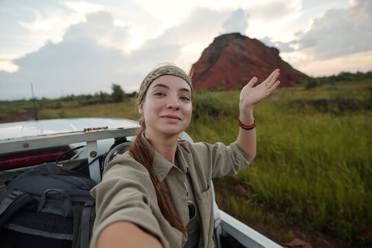 Young satisfied female traveler and blogger looking at camera and waving hand to online audience while recording new video for her vlog