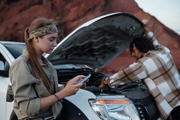 Young woman using mobile application in smartphone while helping her boyfriend bending over engine of pickup truck