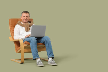 Mature man with laptop messaging in armchair on green background