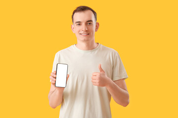 Young man with mobile phone showing thumb-up on yellow background