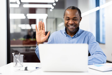 Smiling businessman in office environment engaged video call using laptop. Man waves hand at...