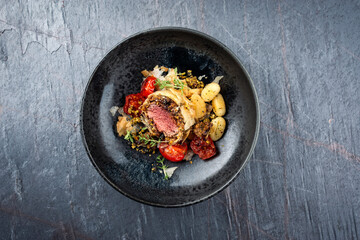 Fried mangalica pork tenderloin coated with crashed pistachio nuts, grated parmesan and chopped cranberries served with tomatoes and gnocchi as top view on a Nordic design bowl