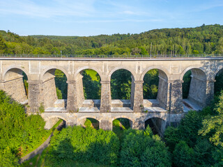 Fototapeta premium The Sychrov Railway Bridge rises gracefully above lush greenery, showcasing its impressive arches against a backdrop of vibrant trees.