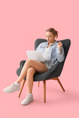 Beautiful young woman with laptop sitting on comfortable armchair against pink background