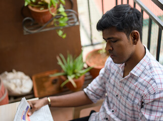 A young man sleeping with newspaper while sitting on a balcony surrounded by potted plants in the morning light