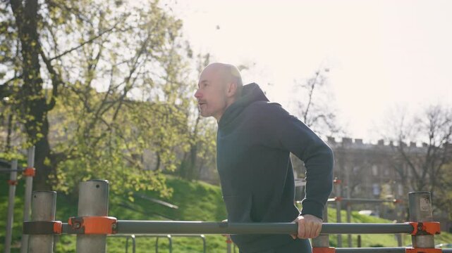 A bald man in a dark hoodie and sweatpants is working out at an urban sports facility. The man is performing dips on parallel bars.