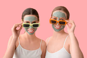 Beautiful young sisters with facial masks and sunglasses on pink background