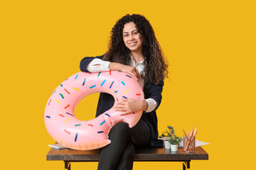 Happy female office worker with inflatable ring sitting on table against yellow background. Summer...