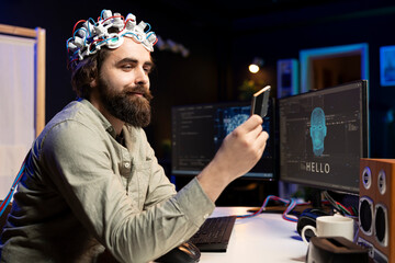 Smiling programmer wearing EEG headset holding disk containing software turning AI sentient,...
