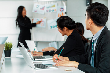 Asian business people listening to presentation from businesswoman who stands in front of group meeting room at international business conference. Multicultural corporate strategy concept. uds