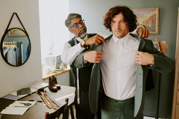 Brutal curly haired model trying on new suit while tailor assisting him