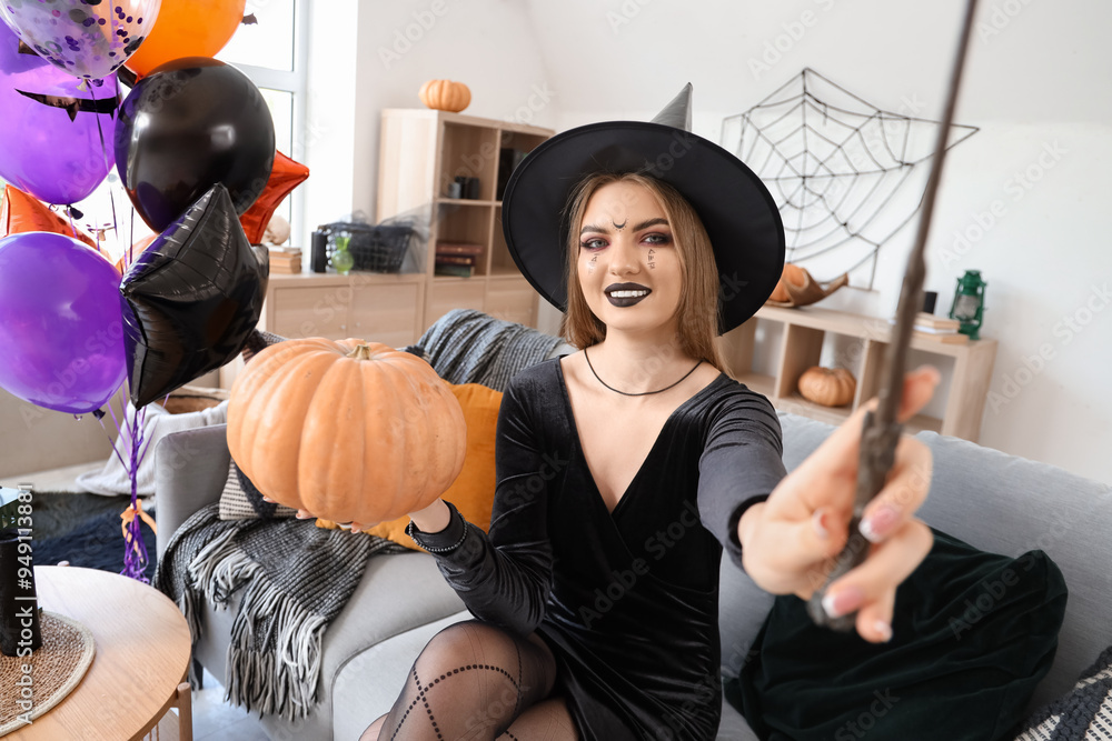 Wall mural Young woman dressed for Halloween as witch with magic wand and pumpkin sitting on sofa at home