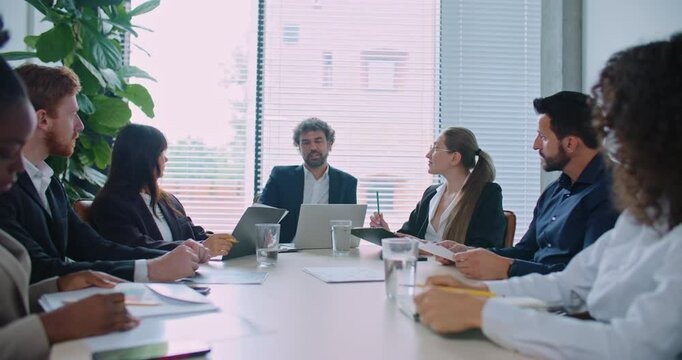 Busy director with his managers sitting at round table in modern room with panoramic window. People having business conference. Discussing about ways of company development. Corporate meeting.
