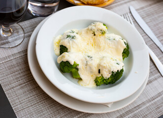 Appetizing broccoli and cauliflower gratin garnished with cheese and cream served on white plate near glass of wine and bread.