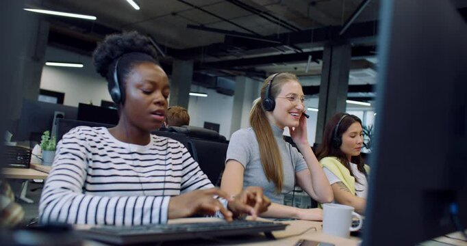 Beautiful diversity workers sitting next to each others at workplace in modern office. Operators of call center answering clients calls. Typing text on keyboards and looking at computers monitor.