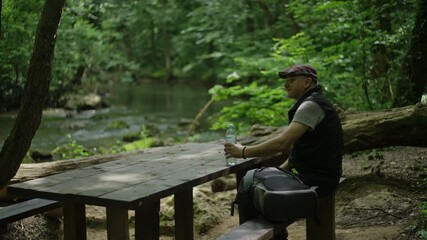 Senior hiker man with backpack resting at a wooden table beside a waterfall and drinking water after a long walk in the forest - Powered by Adobe