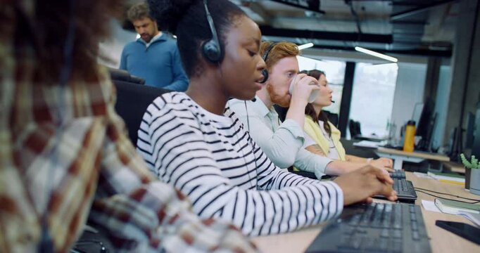 Professional multi-ethnic managers working in call center office. People sitting in row at desk and using computers. Discussing something with customers on headset. Team of workers answering calls.