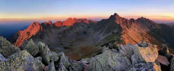 Panoramic view of the Tatra Mountains at sunset, showcasing rugged peaks, craggy cliffs, and dramatic rocky terrain.