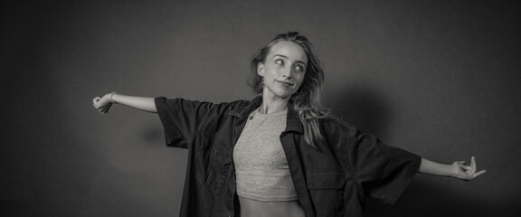 A young woman stands against a dark background with her arms outstretched, wearing a loose shirt and crop top, gazing to the side with a contemplative expression.