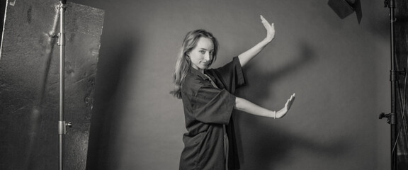 A woman posing with her arms raised in a studio setting, wearing a loose-fitting dark garment, expressing grace and poise in a black and white photograph.