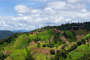 Fototapeta premium Rural mountain village in Northern Thailand. Lush green hills, terraced fields, and traditional houses under a partly cloudy sky.