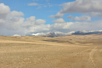 Fototapeta premium Field roads cross high hills in the dry autumn steppe at the foot of high snow-capped mountains.