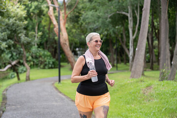 Smiling senior woman jogging with towel and water in forest park