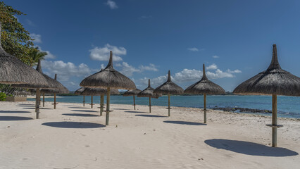 Rows of straw sun umbrellas on the beach. Round shadows on the sand. Turquoise ocean in the distance. Clouds in the blue sky. Mauritius. Belle Mare.
