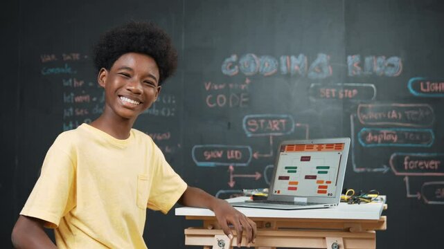 Smart african boy use laptop to code program and turn around to wave hand. Cute high school stident coding engineering prompt or program system at blackboard in STEM technology classroom. Edification