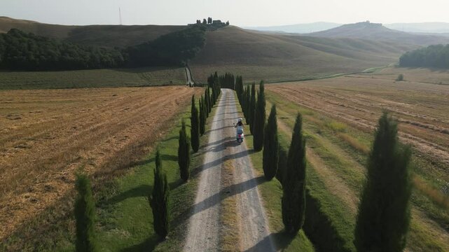 Two people riding a scooter on a scenic road lined with trees through Tuscany in the late afternoon