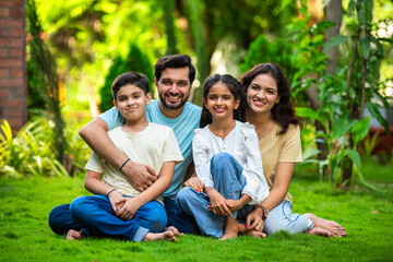 Happy Indian family of four sitting in park, embracing and smiling at camera, enjoying quality time