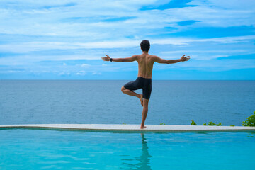 Fototapeta premium Man doing yoga and meditating in tree position near swimming pool on the tropical beach in Samui, Thailand. Healthy lifestyle active balanced practicing meditate and energy, yoga in morning.