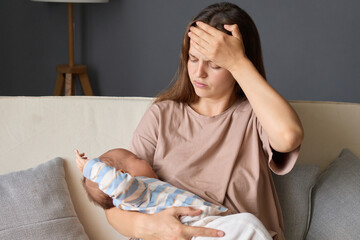 Caucasian tired exhausted woman gently rocking her newborn baby while sitting in couch in the...