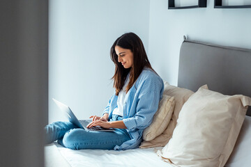 A young millennial woman sits on her bed in a hotel or bedroom, working on her laptop, searching...