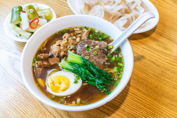 Close-up of Taiwanese Beef Noodle Soup (Niúròu miàn) with Spoon, Dumplings, and Pickles in the Background