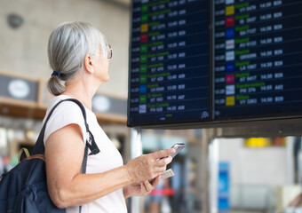 Smiling senior woman in airport looking at timetable schedule to check flight departure gate....