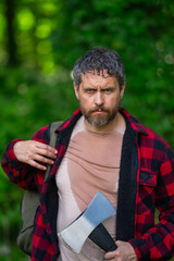 Fototapeta premium Confident lumberjack works in summer forest. Man with an ax. Woodcutter in plaid shirt in forest. Hipster with axe. Lumberjack with an axe. Bearded lumberjack. American lumberjack with hatchet.