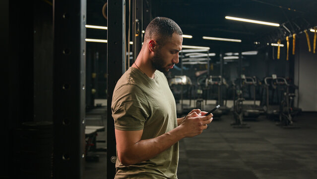 American Latino ethnic man browsing smartphone serious athlete fit sport guy personal fitness trainer instructor male in gym bodybuilder sportsman using mobile phone looking at camera happy smiling