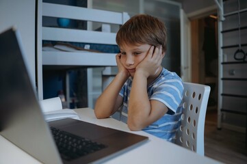 cute little boy studying at home usung laptop, tired, sressed, thinking hard trying to do homework