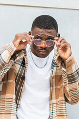 African American man in trendy checkered shirt putting on glasses while leaning on wall on city street