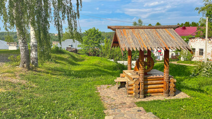 A wooden pavilion sits in a grassy field. The pavilion is surrounded by trees and has a bench underneath it. The scene is peaceful and serene