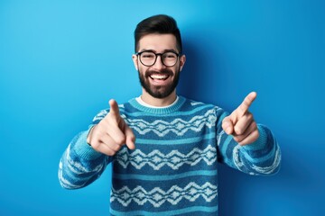 Naklejka premium Handsome bearded man in casual sweater and glasses standing in front of blue background. He smiling and pointing to the camera with fingers, giving a thumbs up.