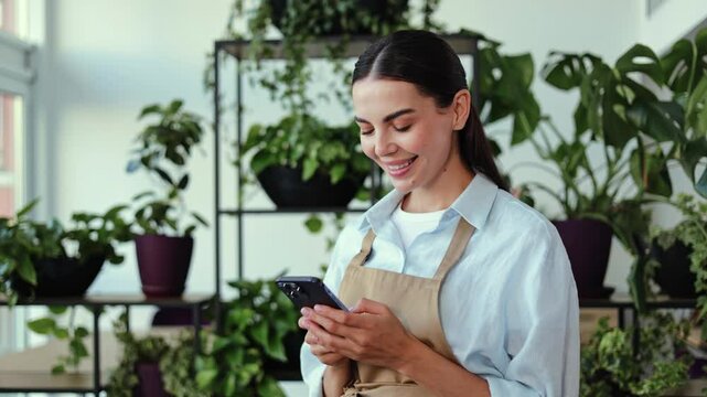 Young woman working at her plant shop. Girl wearing an apron and is smiling while using her phone. She is standing in front of shelves full of potted plants. Natural light shines through the windows.