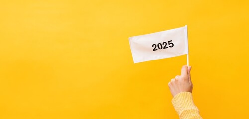 Waving in the Future: Hand Holding a '2025' Flag on Vibrant Yellow Background