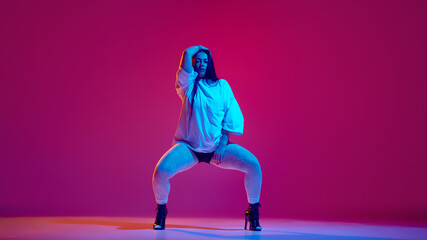 Sensual young woman in oversized t shirt and black heeled shoes dancing high heel dance against magenta studio background. Concept of hobby, entertainment, performance, creativity, lifestyle
