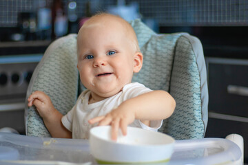 Cute blond caucasian baby child with blue eyes sitting at highchair after eating food. Child is dirty and everything is in rice grains. Baby is serious and surprised