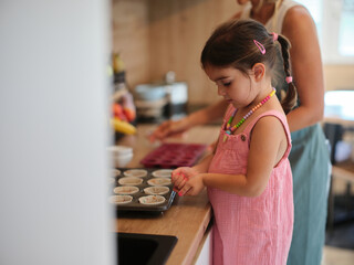 Young girl helps prepare muffin batter in a cozy kitchen alongside an adult, showcasing their...
