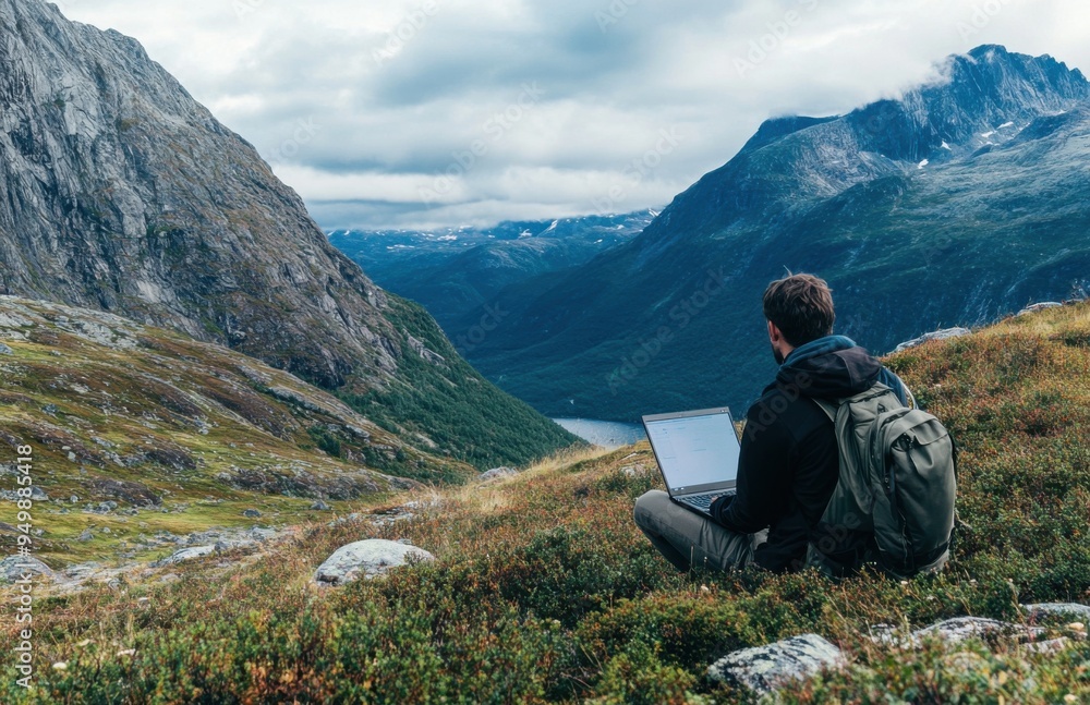 Poster man sits on the grassy ground with his laptop, surrounded by mountains and forests. he gazes at the 