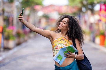 Young woman with curly hair, wearing vibrant summer clothes, holds a map and a smartphone, walking in a colorful, sunny street. She appears focused as she navigates, suggesting a travel or exploration