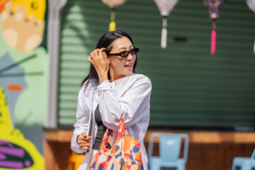 A young Asian woman wearing sunglasses and a white shirt smiles while holding a tablet close to her chest. She walks energetically down a sunlit city street, embodying a modern, tech-savvy lifestyle.